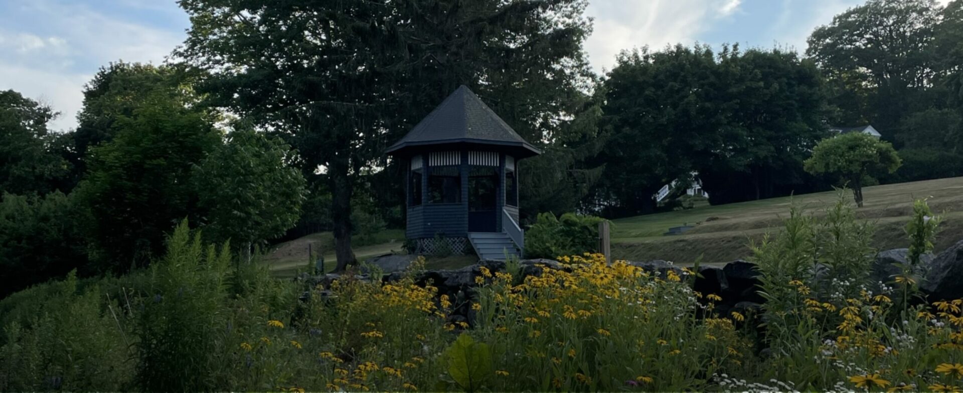 A gazebo in a grassy meadow surrounded by trees and flowers on The Norumbega property